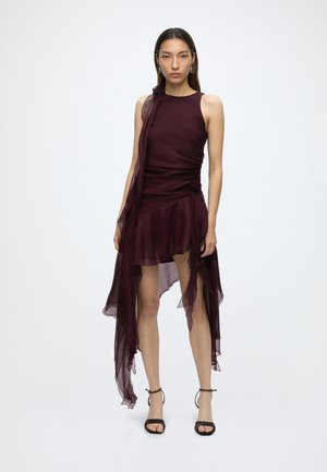 Femme portant une robe asymétrique bordeaux foncé sans manches avec un tissu transparent et des sandales noires à talons hauts, debout devant un fond blanc.
