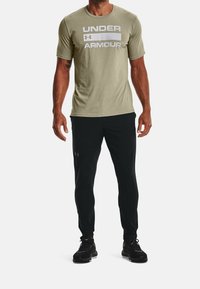 Olivgrön t-shirt med kort ärm och vit "UNDER ARMOUR"-logotyp, ihop med svarta smala byxor och svarta träningsskor. Slät tygtextur.