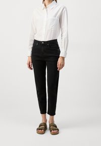 Vit långärmad skjorta med en ficka, kombinerad med svarta slim-fit jeans och olivgröna sandaler med justerbara remmar. Minimalistisk design.