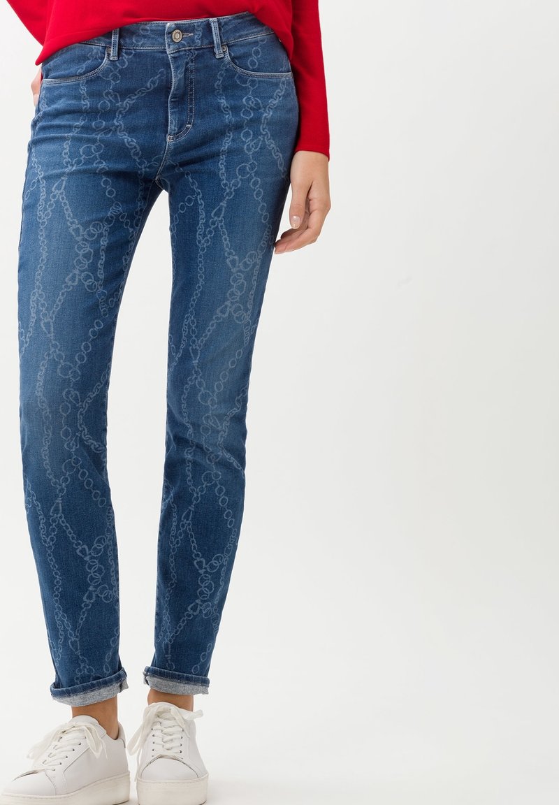 Jean bleu coupe slim avec un motif léger en forme de chaîne, ourlets retroussés, porté avec des baskets blanches et un haut rouge à manches longues.