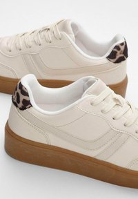 Sneakers beige con una parte superiore sintetica liscia, lacci bianchi e una suola in gomma marrone, con un accento a stampa leopardata sul tallone.