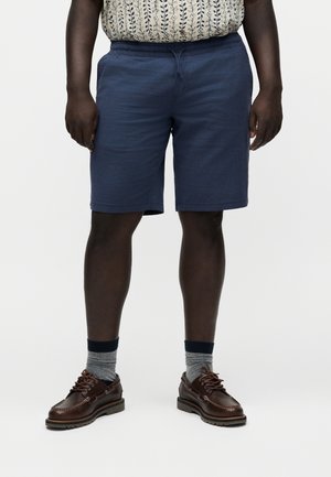Homme portant un short bleu marine, des chaussettes grises avec le haut noir, des chaussures en cuir marron à lacets et une chemise beige à motifs.