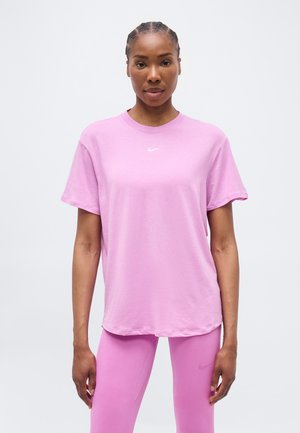 Γυναίκα με πλεξούδες, φορώντας ένα ανοιχτό ροζ μπλουζάκι Nike και ροζ κολάν που ταιριάζει, στέκεται μπροστά σε ένα απλό ανοιχτό φόντο.