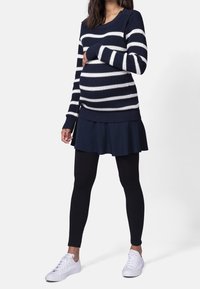 Těhotná žena oblečená v námořnickém a bílém pruhovaném svetru, námořnické sukni, černých legínách a bílých teniskách, stojící proti jednoduchému pozadí.