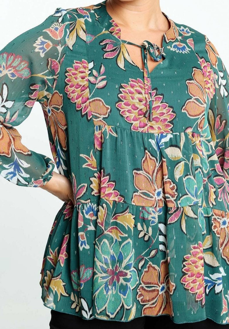 Gros plan sur une personne portant un chemisier vert à fleurs roses, orange, bleues et beiges, avec un lien à nouer au niveau de l'encolure.