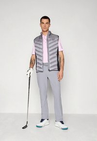 Ανδρική εμφάνιση γκολφ με γκρι αμάνικο μπουφάν, ανοιχτό ροζ πόλο, γκρι παντελόνι και λευκά παπούτσια με μπλε λεπτομέρειες, κρατώντας ένα γκολφ κλαμπ.