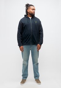 Άντρας με κοτσιδάκια φοράει μαύρο φούτερ με φερμουάρ, ανοιχτό μπλε τζιν και μπεζ αθλητικά, στέκεται μπροστά σε απλό λευκό φόντο.