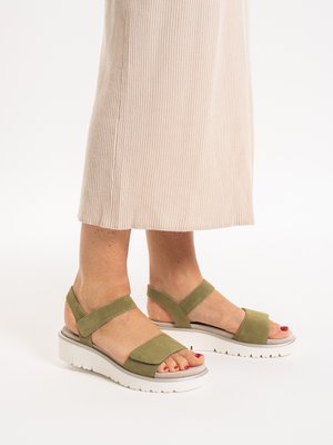 Füße in olivgrünen Sandalen mit Klettverschluss und weißen, dicken Sohlen, getragen zu einem hellbeigen, gerippten Rock vor weißem Hintergrund.