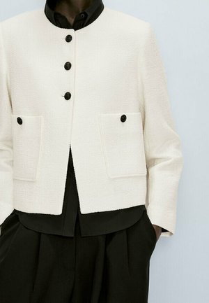 Veste blanche texturée avec des boutons noirs sur une chemise noire et un pantalon noir plissé, portée par une personne les mains dans les poches.