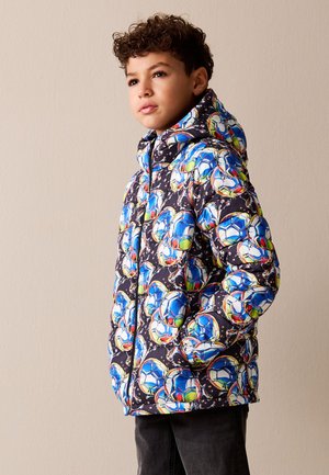 Niño con una chaqueta acolchada con un patrón colorido de balones de fútbol en azul, blanco y verde, de pie con las manos en los bolsillos frente a un fondo liso.