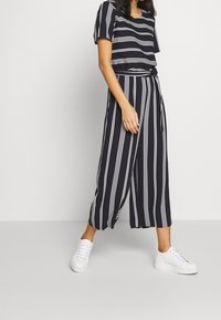Mujer vistiendo pantalones de pierna ancha a rayas blancas y negras y una blusa a juego con zapatillas blancas frente a un fondo gris.