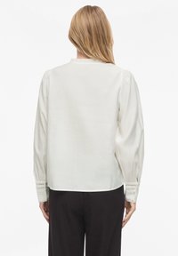 Balta, ilgomis rankovėmis palaidinė iš lygaus audinio. Turi surinktus pečius ir detalias rankogalius su subtilia nėrinių apdaila.