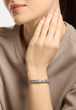 Hand van een vrouw die haar nek aanraakt, ze draagt een beige top en een blauw-gouden armband met vierkante schakels om haar pols.