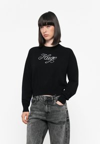 Suéter negro corto con el nombre de la marca "Hugo" bordado en gris claro en el pecho. Combinado con jeans de tiro alto en gris desgastado.
