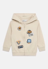 Beige hettejakke med glidelås og hette, dekorert med flere fargerike lapper med racingtema på forsiden, laget av mykt bomullsmateriale.