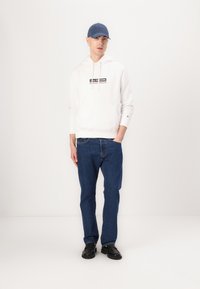 Hvid hættetrøje med et grafisk logo, blå denim jeans og sorte sko, suppleret med en blå kasket. Afslappet pasform, ensfarvede farver.