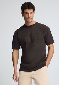 Brun t-shirt med kort ärm och rund halsringning med en tonad "POLO CLUB" grafik på framsidan, i kombination med ljusa byxor.