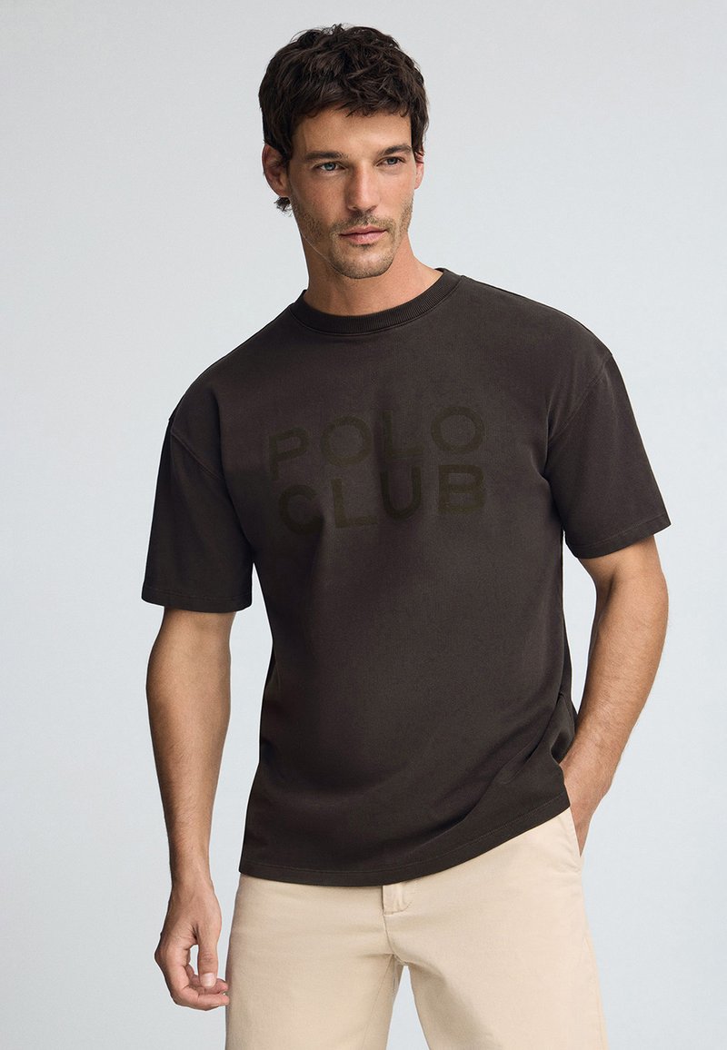 Brun t-shirt med kort ärm och rund halsringning med en tonad "POLO CLUB" grafik på framsidan, i kombination med ljusa byxor.