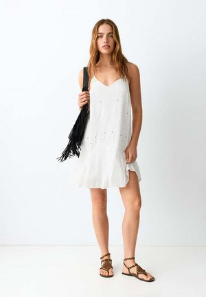 Jeune femme en robe blanche sans manches tenant un sac noir à franges, portant des sandales plates marron à clous, debout devant un fond blanc uni.