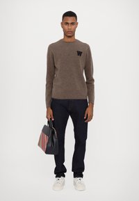 Brown knit sweater with a round neck and black letter "W" logo. Paired with dark jeans, white sneakers, and a multicolored striped handbag.