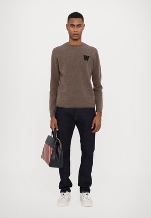 Brown knit sweater with a round neck and black letter "W" logo. Paired with dark jeans, white sneakers, and a multicolored striped handbag.