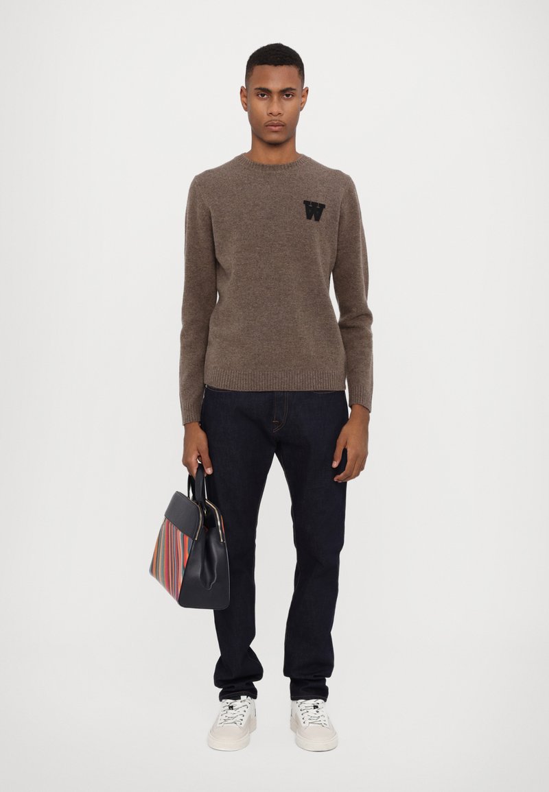 Brown knit sweater with a round neck and black letter "W" logo. Paired with dark jeans, white sneakers, and a multicolored striped handbag.