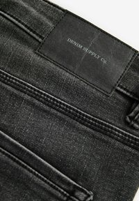 Černá denimová tkanina s hladkým povrchem, obsahující koženou etiketu s vyraženým nápisem "DENIM SUPPLY CO." a viditelnými švy.