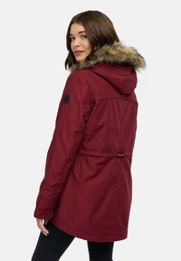 Parka bordeaux con cappuccio in pelliccia sintetica, vita regolabile e dettaglio con bottone nero. Tessuto testurizzato con finitura liscia, design lungo fino al ginocchio.