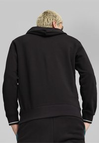 Felpa nera con cappuccio, dotata di polsini e orlo a costine. Presenta una texture morbida. La vista posteriore mostra un design elegante e minimalista.