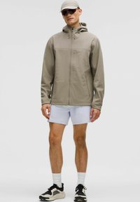 Chaqueta con cremallera gris claro y capucha, con bolsillos laterales, textura suave, llevada sobre una camiseta blanca y pantalones cortos, combinada con zapatos deportivos y una gorra.