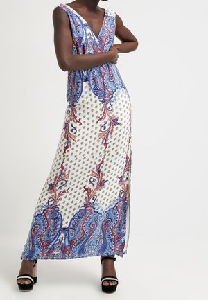 Vrouw die een mouwloze maxi-jurk met V-hals draagt, bedrukt met blauw, rood en wit paisley- en stippenpatronen, gecombineerd met zwarte platformsandalen.