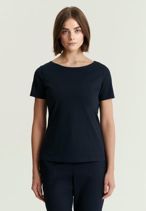 Vrouw met schouderlang bruin haar, die een effen marineblauw T-shirt met korte mouwen en bijpassende broek draagt, staand tegen een lichte achtergrond.