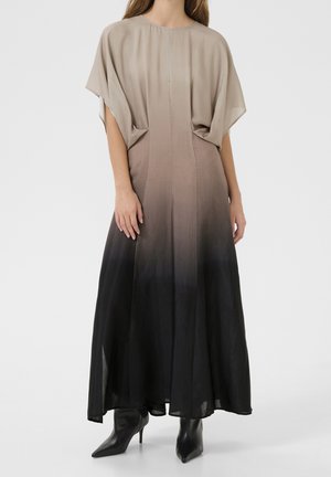 Kvinde iført en lang, løs ombre-kjole, der går fra beige til sort, kombineret med sorte ankelstøvler med spids tå og hæl, stående på hvid baggrund.