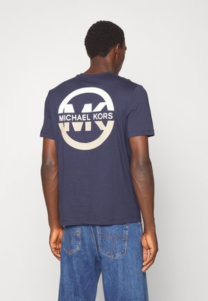 Homme portant un t-shirt bleu marine Michael Kors avec un grand logo circulaire blanc et beige dans le dos, associé à un jean bleu, debout devant un fond blanc.