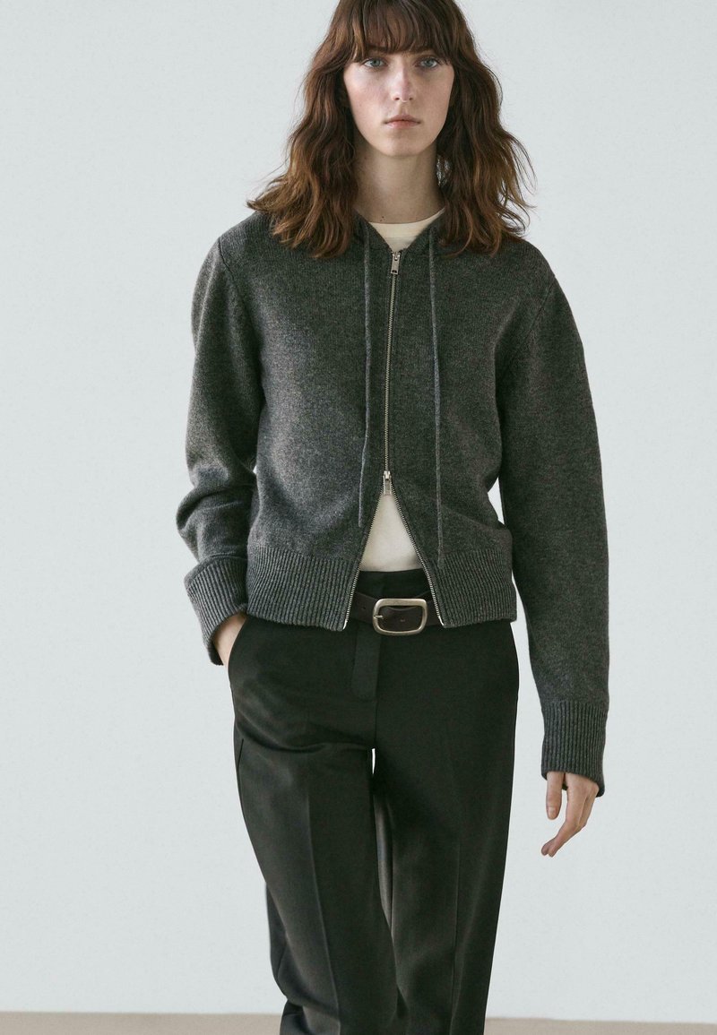 Gray zip-up knit sweater with ribbed cuffs and hem, worn over a white shirt, paired with black tailored trousers. Simple design and textured fabric.