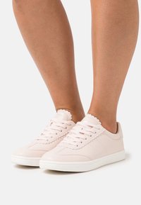 Pieds portant des baskets lacées rose clair avec des bords festonnés et des semelles blanches sur un fond blanc uni.