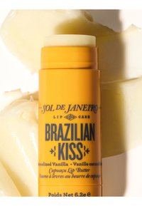 Keltainen huulirasvatuubi, jossa on Brazilian Kiss -etiketti, karamellisoitu vanilja ja cupuaçu-huulevoide, asetettuna vaalealle kermainen teksturoidulle taustalle.