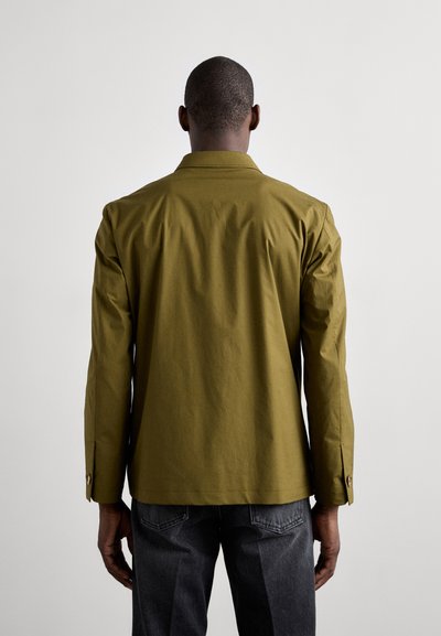 Khaki knap-shirt med lange ærmer, rygventilation og træknapdetaljer. Kombineret med mørkegrå jeans. Glat tekstur.