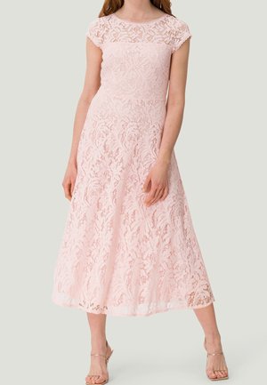 Femme portant une robe midi en dentelle rose pâle avec manches courtes et corsage ajusté, associée à des talons nus à bouts ouverts, debout sur un fond neutre.