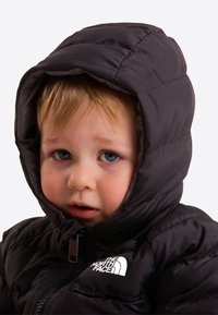Jeune enfant aux cheveux clairs et aux yeux bleus portant une doudoune noire à capuche avec le logo The North Face.