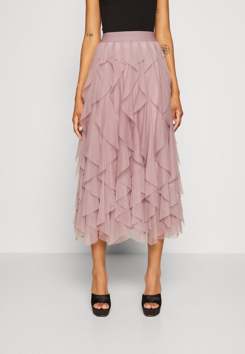 Femme portant une jupe midi rose poudré à couches avec un tissu en tulle froncé et des chaussures noires à talons hauts et bouts ouverts, debout devant un fond uni.