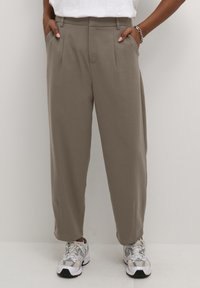 Pantalon plissé taille haute couleur taupe porté avec des baskets blanches et un haut blanc, mains dans les poches, bracelet argenté au poignet droit.