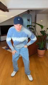 Light blue and white striped fuzzy sweater, blue jeans, and white sneakers; standing with hands on knees in a living room setting.