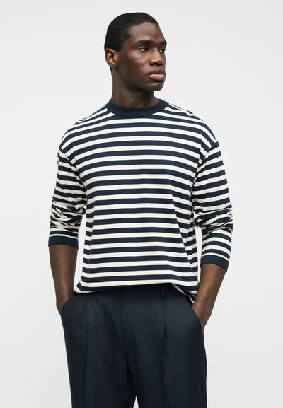 Άνδρας που φοράει μακρυμάνικο πουκάμισο με οριζόντιες ρίγες σε ναυτικό μπλε και λευκό και σκούρο ναυτικό μπλε παντελόνι, στέκεται με τα χέρια στις τσέπες, κοιτάζοντας προς τα πλευρά.