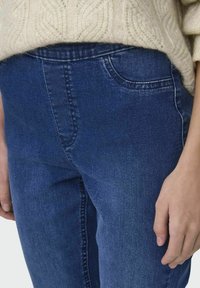 Jeans de mezclilla azul oscuro con una textura suave, diseño de cinco bolsillos y costuras cosidas, combinados con un suéter de punto trenzado color crema.