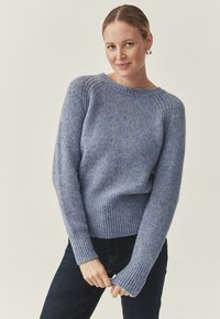 Pull en tricot bleu avec un col rond et des poignets côtelés. Texture douce, coupe ample et manches longues. Porté avec un jean foncé.