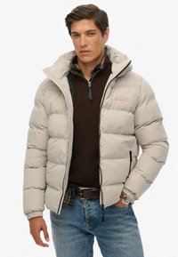 Chaqueta acolchada gris claro con cierre frontal, que presenta una capucha y un logo en el pecho. Llevada sobre un suéter marrón oscuro y unos jeans azules.