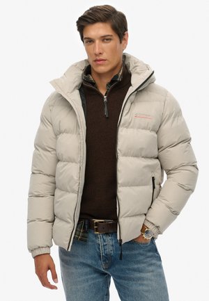 Chaqueta acolchada gris claro con cierre frontal, que presenta una capucha y un logo en el pecho. Llevada sobre un suéter marrón oscuro y unos jeans azules.