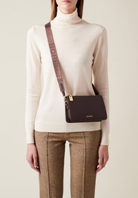 Borsa a spalla in pelle marrone con hardware dorato e una larga tracolla in tessuto. Sono visibili anche pantaloni beige testurizzati e un maglione a dolcevita color crema.