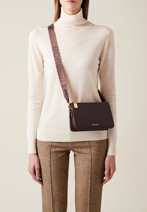 Sac en cuir marron avec des ferrures dorées et une large sangle en tissu. Un pantalon beige texturé et un pull col roulé crème sont également visibles.